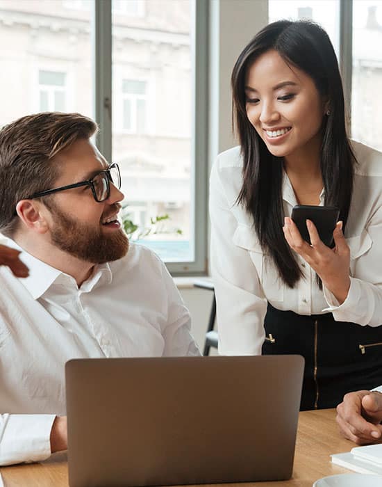 Professional team collaboration in a modern office, featuring a smiling woman holding a smartphone and a man engaging with a laptop, representing PMGS Digital Marketing's focus on digital solutions and client interaction.
